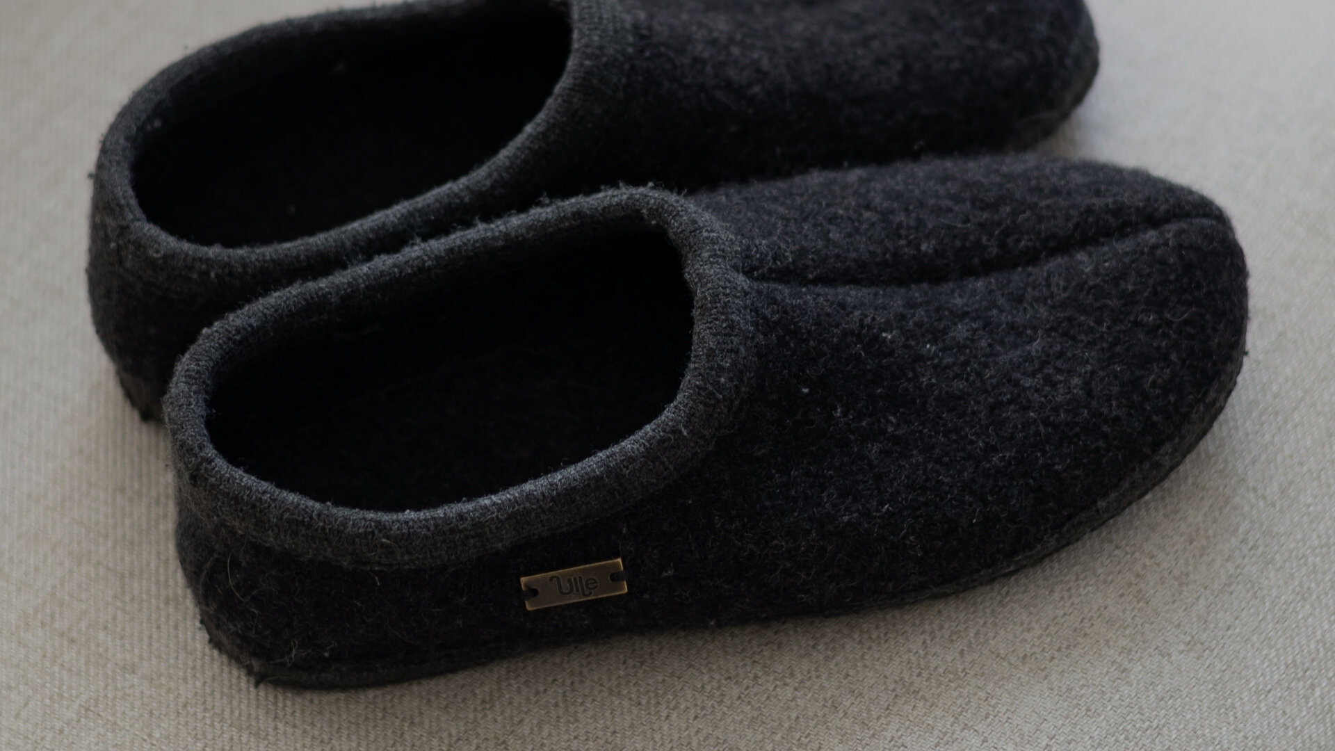 Mörkgrå Ulletofflor i mjuk ull, stilfullt placerade på en ljus soffa – en varm och tidlös favorit för Black Week 2025.